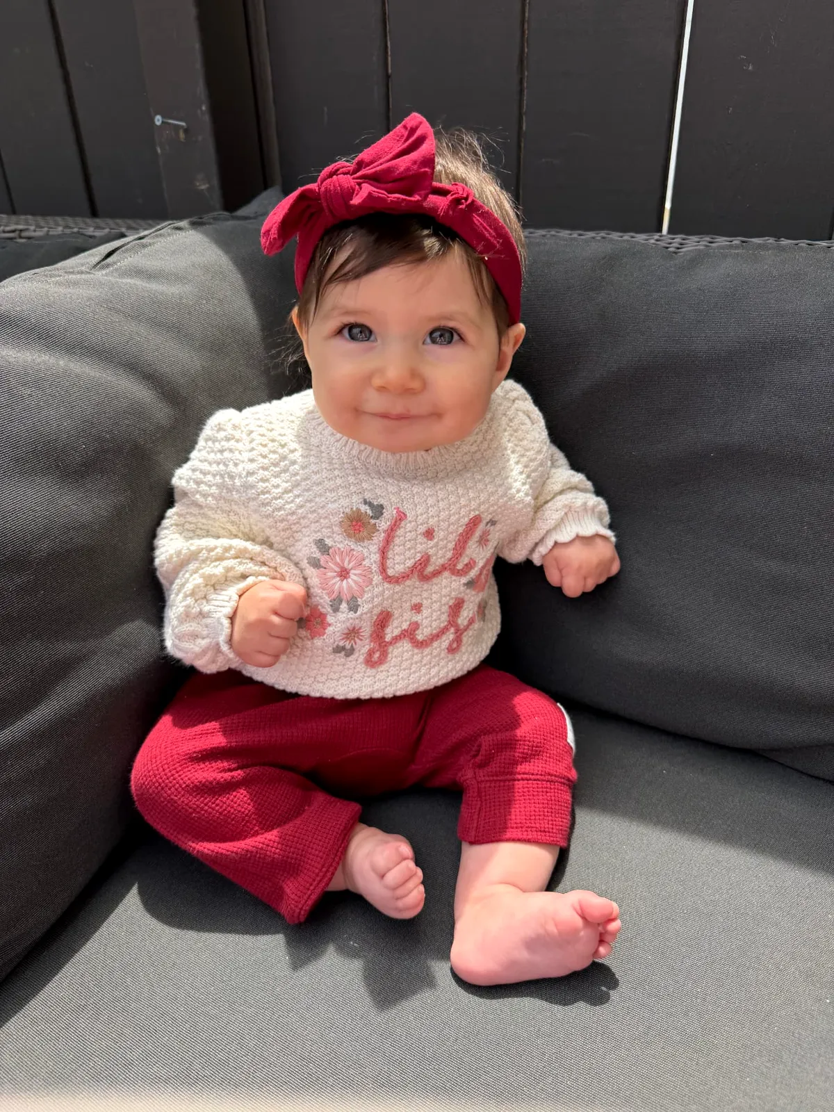 An older baby in a red bow and sweater sitting up on the couch